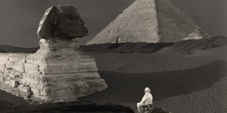 A view of the Sphinx still covered in sand. Behind it, the Great Pyramid.