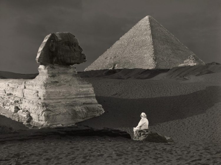 A view of the Sphinx still covered in sand. Behind it, the Great Pyramid.