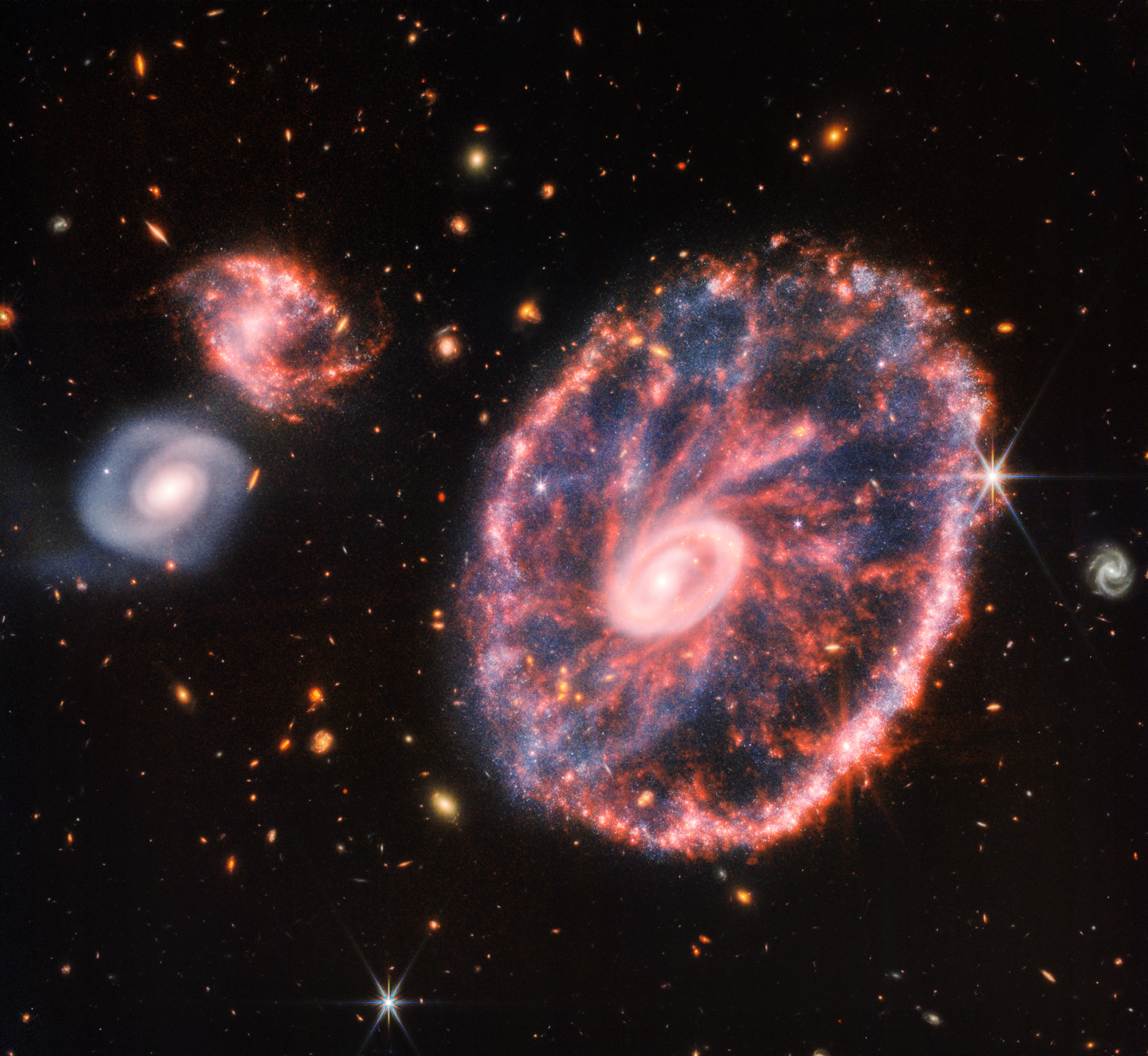The right side of the galaxy is a large pink, speckled wheel with an inner oval, with dusty blue in between, and the left side is two spiral galaxies about the same size. Credits: NASA, ESA, CSA, STScI.