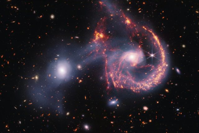 This striking image of Arp 107, captured by the James Webb Space Telescope’s NIRCam and MIRI instruments, offers a detailed look at the aftermath of a galactic collision. The near-infrared data (white) highlights older stars and the faint bridge of gas and stars linking the two galaxies. Meanwhile, the mid-infrared view (orange-red) focuses on younger stars and active star-forming regions. The collision, which occurred hundreds of millions of years ago, triggered new star formation and left the spiral galaxy with a distinctive “smile.” Credit: NASA, ESA, CSA, STScI.