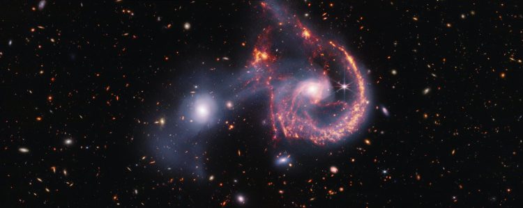 This striking image of Arp 107, captured by the James Webb Space Telescope’s NIRCam and MIRI instruments, offers a detailed look at the aftermath of a galactic collision. The near-infrared data (white) highlights older stars and the faint bridge of gas and stars linking the two galaxies. Meanwhile, the mid-infrared view (orange-red) focuses on younger stars and active star-forming regions. The collision, which occurred hundreds of millions of years ago, triggered new star formation and left the spiral galaxy with a distinctive “smile.” Credit: NASA, ESA, CSA, STScI.