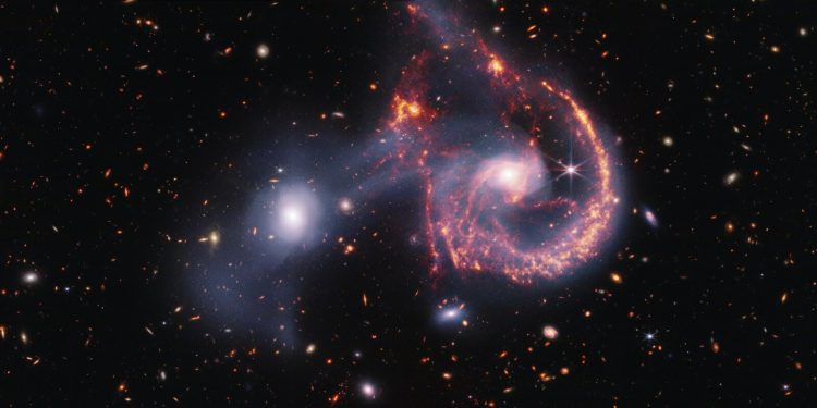 This striking image of Arp 107, captured by the James Webb Space Telescope’s NIRCam and MIRI instruments, offers a detailed look at the aftermath of a galactic collision. The near-infrared data (white) highlights older stars and the faint bridge of gas and stars linking the two galaxies. Meanwhile, the mid-infrared view (orange-red) focuses on younger stars and active star-forming regions. The collision, which occurred hundreds of millions of years ago, triggered new star formation and left the spiral galaxy with a distinctive “smile.” Credit: NASA, ESA, CSA, STScI.