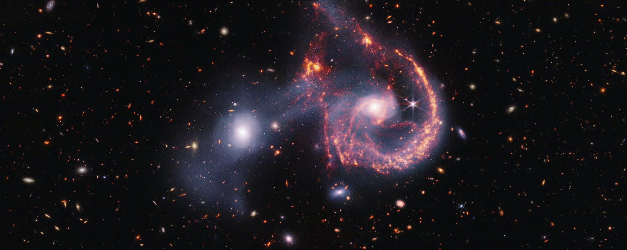 This striking image of Arp 107, captured by the James Webb Space Telescope’s NIRCam and MIRI instruments, offers a detailed look at the aftermath of a galactic collision. The near-infrared data (white) highlights older stars and the faint bridge of gas and stars linking the two galaxies. Meanwhile, the mid-infrared view (orange-red) focuses on younger stars and active star-forming regions. The collision, which occurred hundreds of millions of years ago, triggered new star formation and left the spiral galaxy with a distinctive “smile.” Credit: NASA, ESA, CSA, STScI.