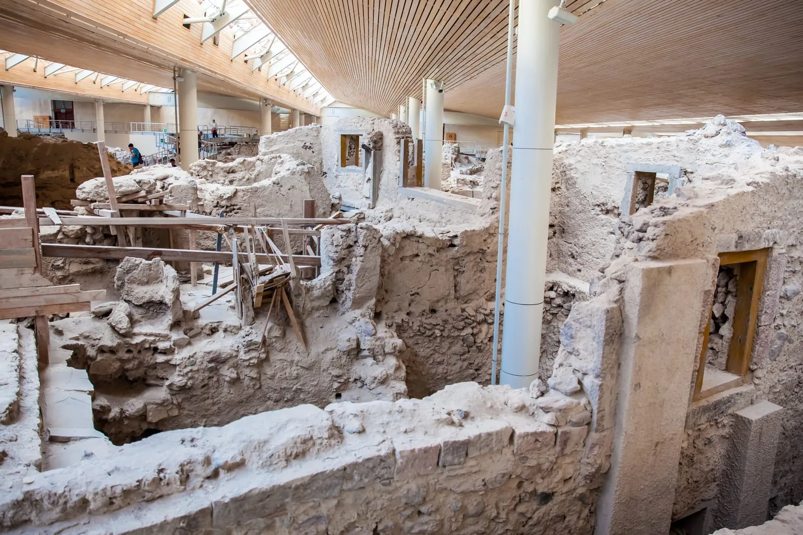 Inside the covered archaeological site, visitors walk past the preserved ruins of Akrotiri. The structure in view likely formed part of a multi-story residence, with surviving windows and entryways still visible.