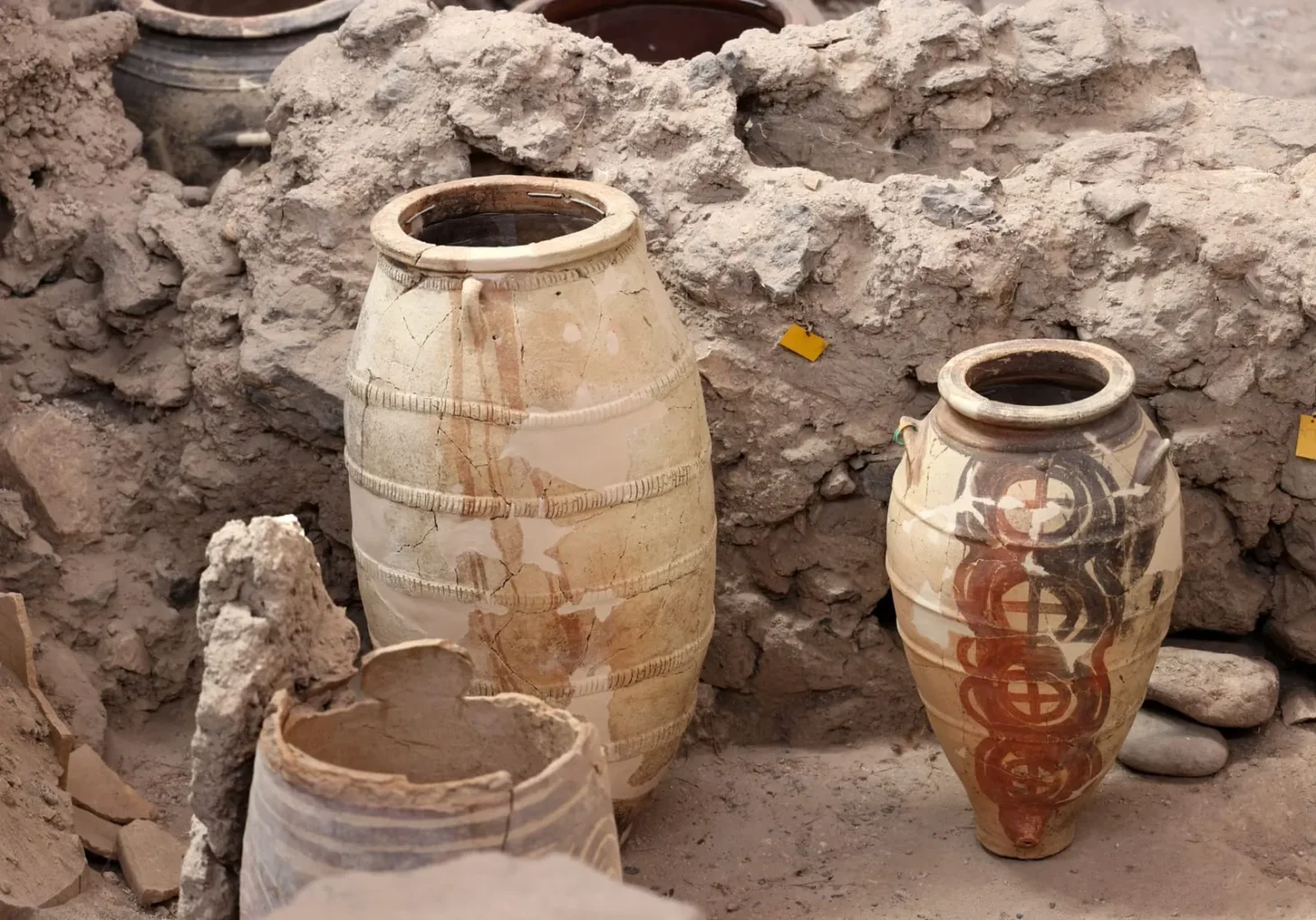 Two large Minoan storage jars stand intact amid the ruins of Akrotiri. The vessel on the right is decorated with circular red and black motifs, while the one on the left appears restored and patched. Their preservation shows how well the volcanic ash sealed the site.