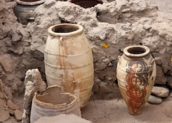 Two large Minoan storage jars stand intact amid the ruins of Akrotiri. The vessel on the right is decorated with circular red and black motifs, while the one on the left appears restored and patched. Their preservation shows how well the volcanic ash sealed the site.