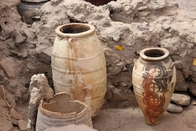 Two large Minoan storage jars stand intact amid the ruins of Akrotiri. The vessel on the right is decorated with circular red and black motifs, while the one on the left appears restored and patched. Their preservation shows how well the volcanic ash sealed the site.